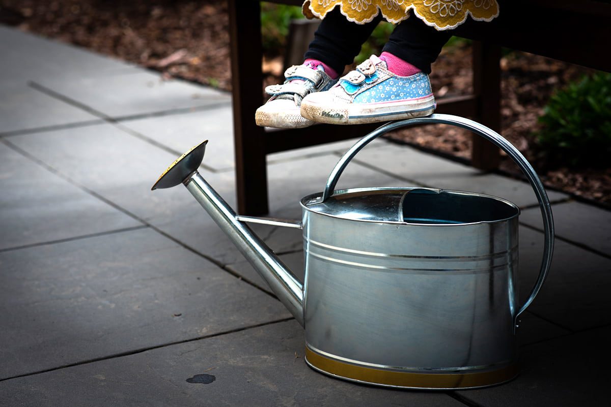 Child's feet dangling off of a bench with a watering can in the foreground.